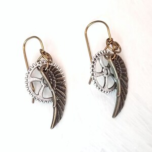 Peut inclure: Une paire de boucles d'oreilles en argent et en laiton avec un charme de roue dentée et une plume. Les boucles d'oreilles sont sur un fond blanc.
