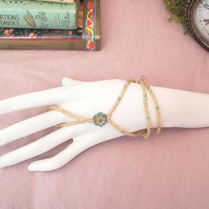 May include: A gold and turquoise beaded bracelet with a flower charm. The bracelet is on a white hand model.