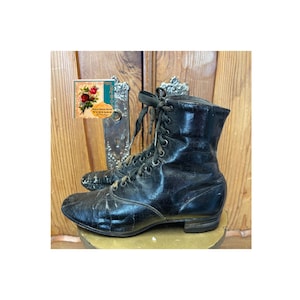 May include: A pair of vintage black leather lace-up boots. The boots have a worn, distressed appearance, suggesting age and use. The laces are dark, and the boots sit on a round, golden-colored surface. A decorative tag with floral imagery is visible.