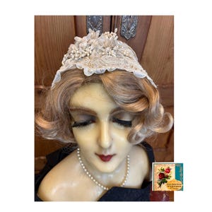 May include: A vintage-style cream-colored lace headpiece with floral embellishments. The headpiece is displayed on a mannequin head with blonde hair, a pearl necklace, and a black top. A small vintage label is visible.