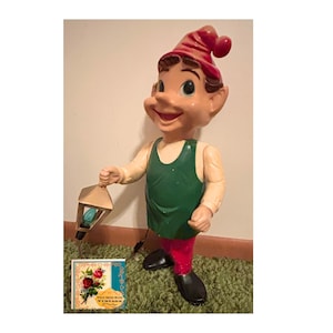 May include: A molded plastic elf figurine wearing a red pointed hat, a green sleeveless tunic, and red pants with black shoes. The elf holds a small, metallic-colored lantern with a blue light bulb inside. A square tag with floral art reads Wild Rose Vintage.
