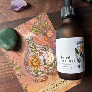 May include: A brown glass bottle with a dropper top and a label that reads "Earth Bound Grounding and Calming herbal supplement OZ/ ML FLOWER MOOD" The bottle is sitting on top of a card with an illustration of a plant and the text "Ashwagandha Purpose -54- ".