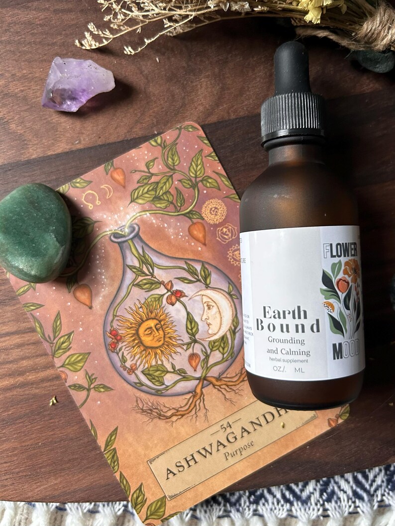 May include: A brown glass bottle with a dropper top and a label that reads "Earth Bound Grounding and Calming herbal supplement OZ/ ML FLOWER MOOD" The bottle is sitting on top of a card with an illustration of a plant and the text "Ashwagandha Purpose -54- ".