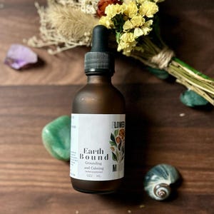 May include: A brown glass bottle with a dropper top and a white label that reads "Earth Bound Grounding and Calming Herbal Supplement 0.2 fl oz / 6 ml". The bottle is sitting on a wooden surface with dried flowers, a purple crystal, a green stone, and a seashell.