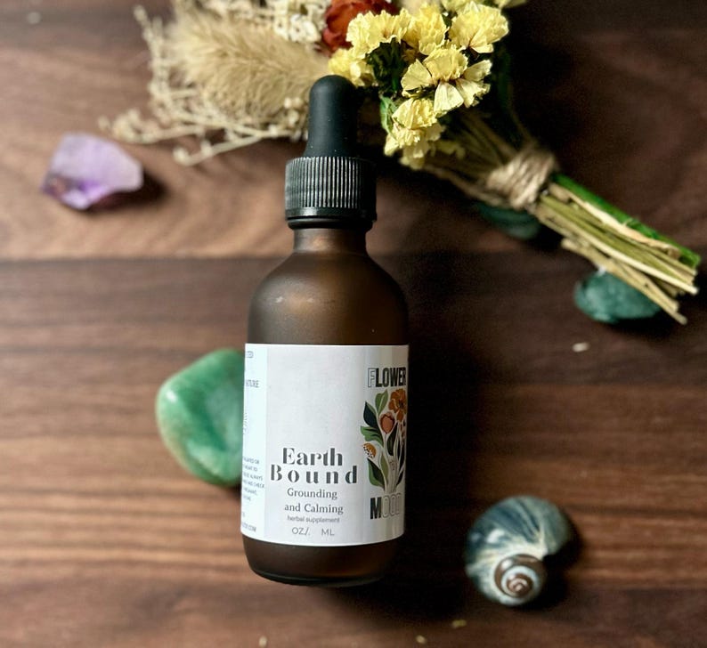 May include: A brown glass bottle with a dropper top and a white label that reads "Earth Bound Grounding and Calming Herbal Supplement 0.2 fl oz / 6 ml". The bottle is sitting on a wooden surface with dried flowers, a purple crystal, a green stone, and a seashell.