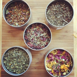 May include: Five round metal tins filled with different types of loose leaf tea. The tins are arranged in a circle on a wooden surface. The teas include a brown tea, a green tea, a red tea, a yellow tea, and a purple tea.