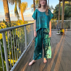 Pode incluir: Um conjunto de pijama tie-dye azul e verde. A parte de cima é de manga curta e curta. As calças são de perna larga com cintura com cordão. O conjunto é feito de material sedoso. A pessoa está em pé numa varanda de madeira.