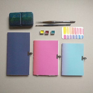 Può includere: Un assortimento di materiali per l'arte ad acquerello, tra cui tre piccoli quaderni blu, rosa e azzurri, una tavolozza per acquerelli, pennelli e una scatola per acquerelli con un motivo a stelle.