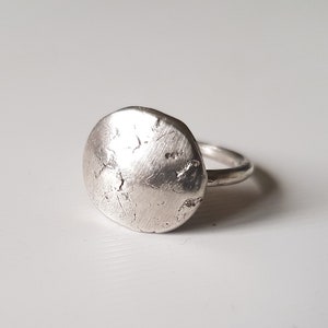 Op de afbeelding: Een zilveren ring met een getextureerde, cirkelvormige voorkant. De band van de ring is dun en glad, in contrast met het ruwe, ongelijke oppervlak van de voorkant. De ring is waarschijnlijk gemaakt van sterling zilver en is een sieraad.