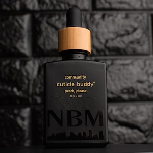 Puede incluir: Una botella negra de aceite para cutículas con tapón y cuentagotas de madera. La botella tiene texto dorado que dice "community cuticle buddy peach, please". La parte inferior de la botella tiene las letras "NBM" y un horizonte urbano.