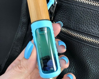 Puede incluir: Un frasco de vidrio azul y transparente con una tapa de madera. El frasco está etiquetado "NBM cuticle buddy 8ml" y contiene piedras azules y verdes.