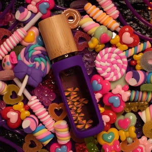 Puede incluir: Un frasco de perfume morado con tapa de madera se encuentra entre una colorida variedad de caramelos, incluyendo piruletas, corazones y gominolas. El frasco está dentro de una funda morada, creando una escena vibrante y juguetona.