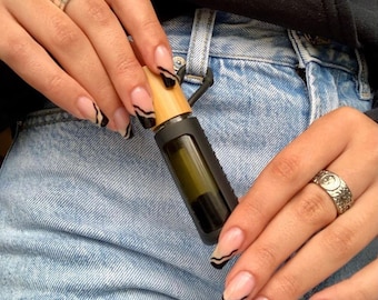 Puede incluir: Primer plano de las manos de una persona con uñas onduladas en blanco y negro. La persona sostiene un pequeño frasco de vidrio verde oscuro con tapa de bambú. El frasco es un frasco de aceite esencial de rodillo.