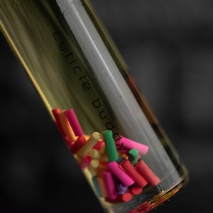 May include: A glass bottle containing a yellow liquid and a collection of colourful, cylindrical objects. The bottle is labelled "cuticle buddy". The cylinders are in shades of pink, orange, yellow, and green. The bottle is clear.