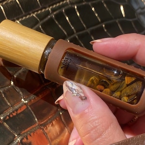以下が含まれることがあります： 木製のキャップと茶色のシリコン製のカバーが付いた茶色のガラス瓶。瓶の中には透明な液体と小さな石が入っています。瓶のラベルには「NBM Beauty Cuticle Buddy」と書いてあります。
