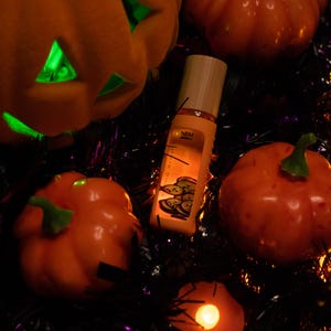Puede incluir: Una botella naranja con tapa de madera está rodeada de calabazas naranjas y una linterna de Jack-o'-lantern tallada. La botella tiene una etiqueta con un gráfico de rodajas verdes. La escena está iluminada con luces naranjas y verdes.
