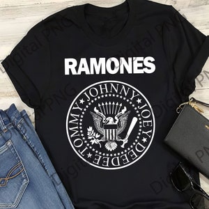 Puede incluir: Camiseta negra con la palabra "RAMONES" en letras blancas sobre un emblema circular. El emblema presenta un águila, estrellas y los nombres "TOMMY", "JOHNNY" y "JOEY".