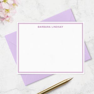 May include: A white stationery card with a purple border and the name "BARBARA LINDSAY" printed at the top. The card rests on a lavender envelope, with a gold pen in the foreground. Pink flowers are in the upper left corner.