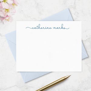 May include: A white rectangular stationery card featuring the name catherine marks printed in a dark blue cursive script font, positioned above a light blue envelope and a gold-toned pen on a white marble surface.