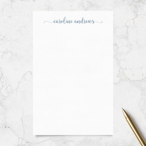 May include: A rectangular white notepad featuring the name caroline andrews printed in a dark blue cursive script at the top center. The notepad is positioned next to a gold-colored pen, providing a clean and simple surface for writing notes.