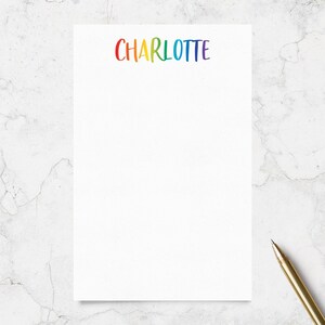 May include: A rectangular white notepad featuring the name CHARLOTTE printed in a vibrant rainbow gradient at the top center. The letters are written in a playful, hand-lettered font style. A gold-toned pen is positioned at the bottom right corner.