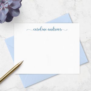 May include: A white stationery card with the name "caroline andrews" in navy blue script, resting on a light blue envelope. A gold pen is in the lower left corner, and a purple succulent is in the upper left corner.