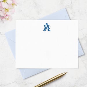 May include: White stationery with a blue monogram, resting on a light blue envelope. A gold pen is positioned near the stationery. Pink flowers are in the upper left corner. The monogram is in a decorative font.
