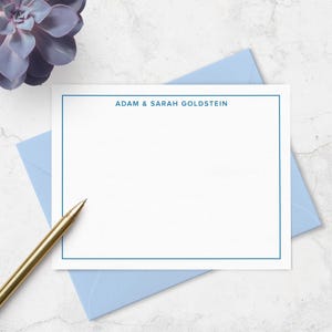 May include: A white rectangular stationery card with a thin blue border and the text ADAM & SARAH GOLDSTEIN printed in blue at the top. The card rests on a light blue envelope, accompanied by a gold pen and a purple succulent plant.