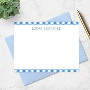 May include: A white rectangular stationery card featuring a blue and white gingham check border at the top and bottom edges. The name LUCAS BRADFORD is printed in a blue sans-serif font in the upper center of the card, resting on a light blue envelope.