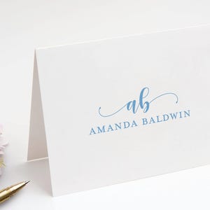 May include: A white folded note card featuring the blue script initials ab above the printed name AMANDA BALDWIN in a serif font. The card is positioned next to a gold pen and a small cluster of light pink flowers with yellow centers.
