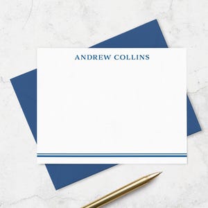 May include: A white rectangular stationery card featuring the name ANDREW COLLINS printed in blue at the top, with two horizontal blue lines at the bottom, accompanied by a matching blue envelope and a gold-toned metal pen.