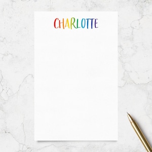 May include: A rectangular white notepad featuring the name CHARLOTTE printed in a rainbow-colored gradient font at the top center. The letters transition through red, orange, yellow, green, blue, and purple hues. A gold pen rests near the bottom right.