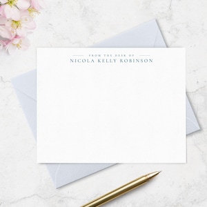 May include: A white rectangular stationery card featuring the dark blue printed text FROM THE DESK OF NICOLA KELLY ROBINSON centered at the top, accompanied by a light blue envelope and a gold-toned metal pen resting on the surface.