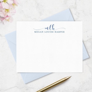 May include: A white rectangular stationery card featuring the dark blue printed monogram mlh in a cursive script above the name MEGAN LOUISE HARPER in a clean sans-serif font, positioned on a light blue envelope with a gold-toned pen nearby.