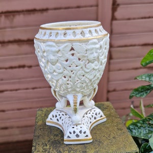 May include: An elegant white vase with gold detailing and a pierced design. The vase has a wide opening and sits on a decorative base. The intricate pattern and gold accents give it a classic, ornamental appearance, suitable for display.