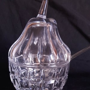 Può includere: Una zuccheriera di vetro trasparente a forma di pera con un coperchio e un motivo decorativo in vetro tagliato. La zuccheriera è su uno sfondo nero.