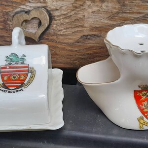 May include: Two vintage white ceramic shaving items. One is a lidded butter dish with a crest that reads "Eastbourne". The other is a shaving scuttle with a handle and a crest that reads "Hastings". Both have gold trim.