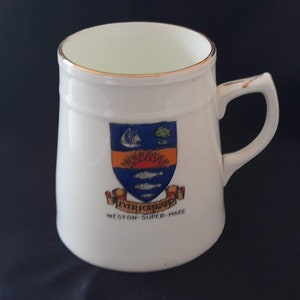 May include: White ceramic mug with a gold rim and a colorful crest featuring a ship, sun, fish, and the text "Ever Forward Weston-Super-Mare".