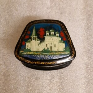 May include: A small, black lacquered wooden box with a hinged lid. The lid is decorated with a painted scene of a white church with red and green trees against a blue sky. The box has gold trim around the edges.