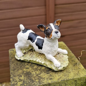 May include: A white and black figurine of a dog with brown markings on its face. The dog is lying down on a green base with a textured surface.