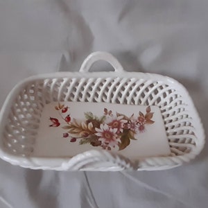 May include: White ceramic basket with a woven design and a floral print on the bottom. The basket has a handle on top.