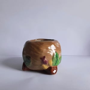 May include: A small, brown ceramic planter with a floral design. The planter has three small feet and is decorated with green leaves and red and yellow flowers.