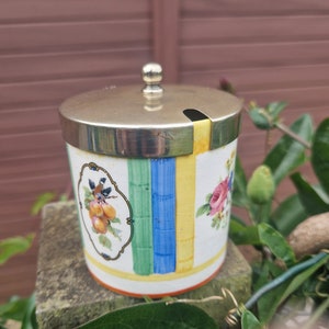 May include: A white ceramic jar with a silver lid and a colorful floral and fruit design. The jar has a spout on the lid and a knob on top.
