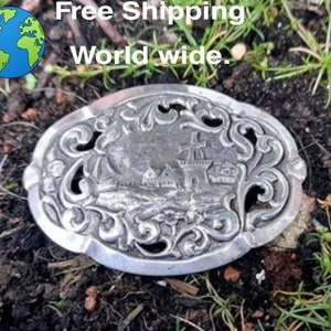 May include: A silver-toned metal belt buckle with an ornate design featuring a windmill and a small village scene.
