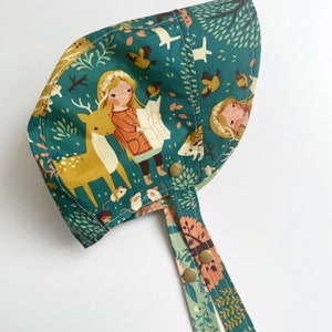 May include: A green fabric sun hat with a woodland print featuring a deer, a girl, and small animals. The hat has two long ties with the same print.
