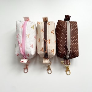 Peut inclure: Trois petites pochettes rectangulaires en tissu avec fermetures éclair et porte-clés. Les pochettes sont blanches, beiges et marron, chacune avec un motif différent de nœuds ou de pois. Les fermetures éclair et les porte-clés sont de couleurs différentes.