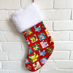 May include: Red Christmas stocking with a white fluffy cuff. The stocking features a repeating pattern of colourful cartoon characters. The stocking is suspended against a white brick wall.