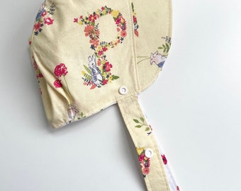 May include: A yellow sun hat with a floral print and a white button closure. The hat has a tie that fastens with buttons. The hat has a floral pattern with a bunny and a pig.