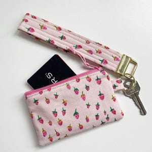 May include: A pink coin purse and wrist strap, both decorated with a strawberry pattern. The purse has a zip closure and a black card inside. A key and gold-coloured clasp are attached to the wrist strap.