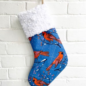 May include: A blue Christmas stocking with a white faux fur cuff. The stocking features a repeating pattern of red cardinals and pink berries on a blue background.
